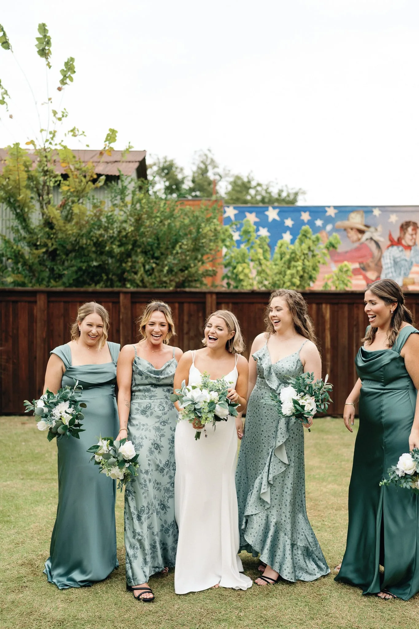 Bride laughing with bridesmaids in sage green before a western mural at a backyard wedding