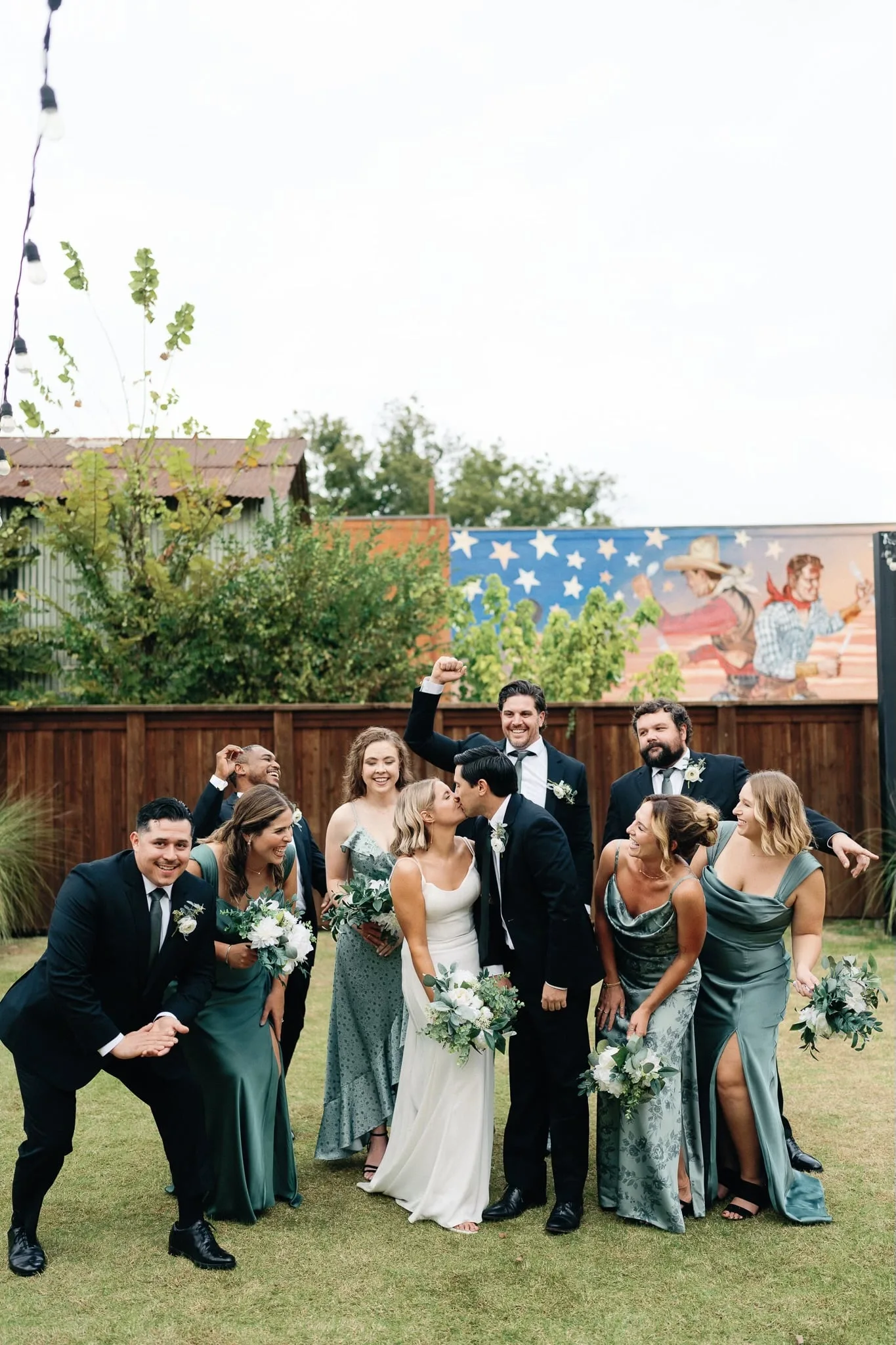 Wedding party celebrating around the kissing newlyweds in a Dallas backyard with cowboy mural