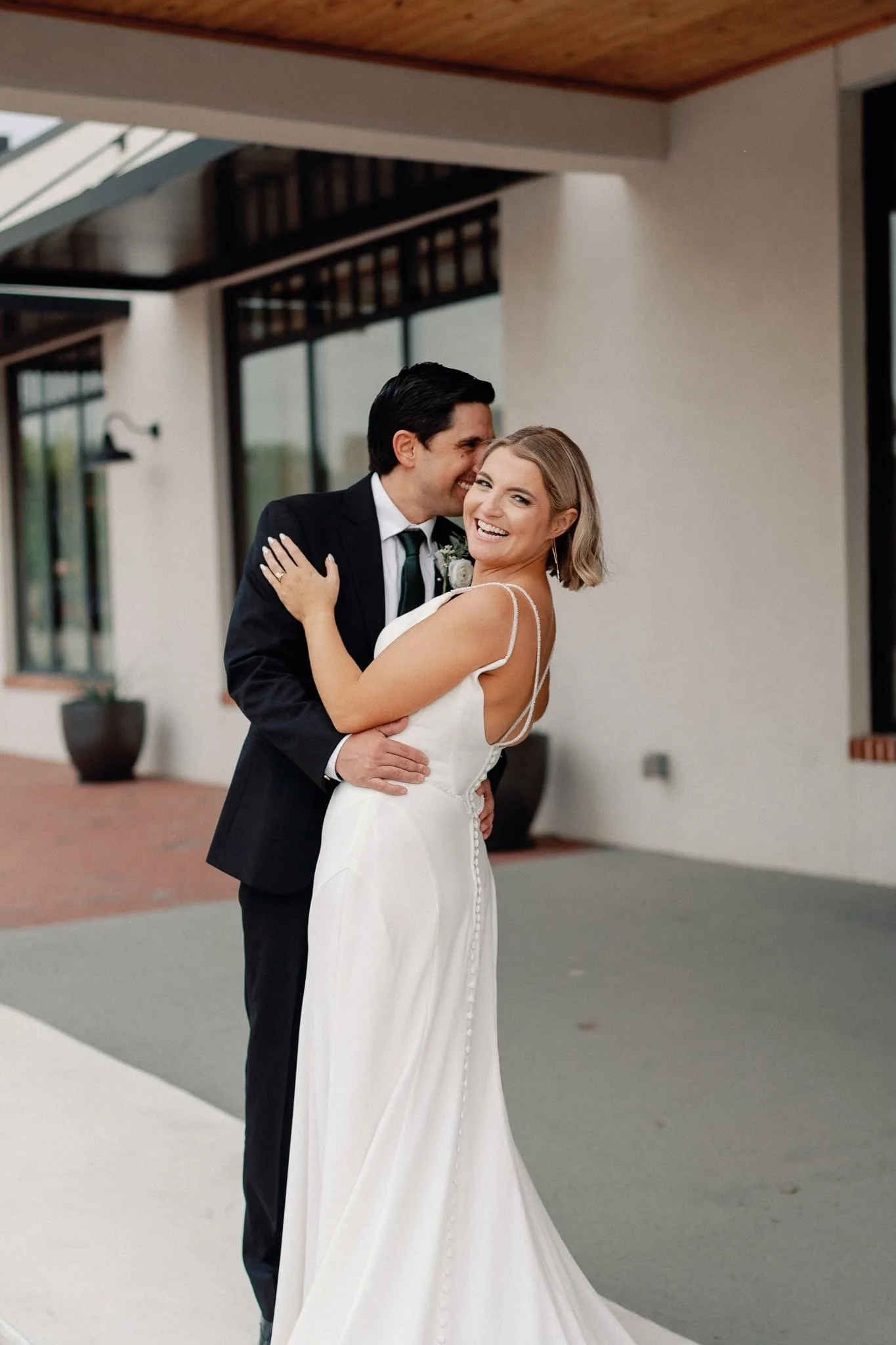 Groom embracing his bride from behind as she smiles under a modern breezeway