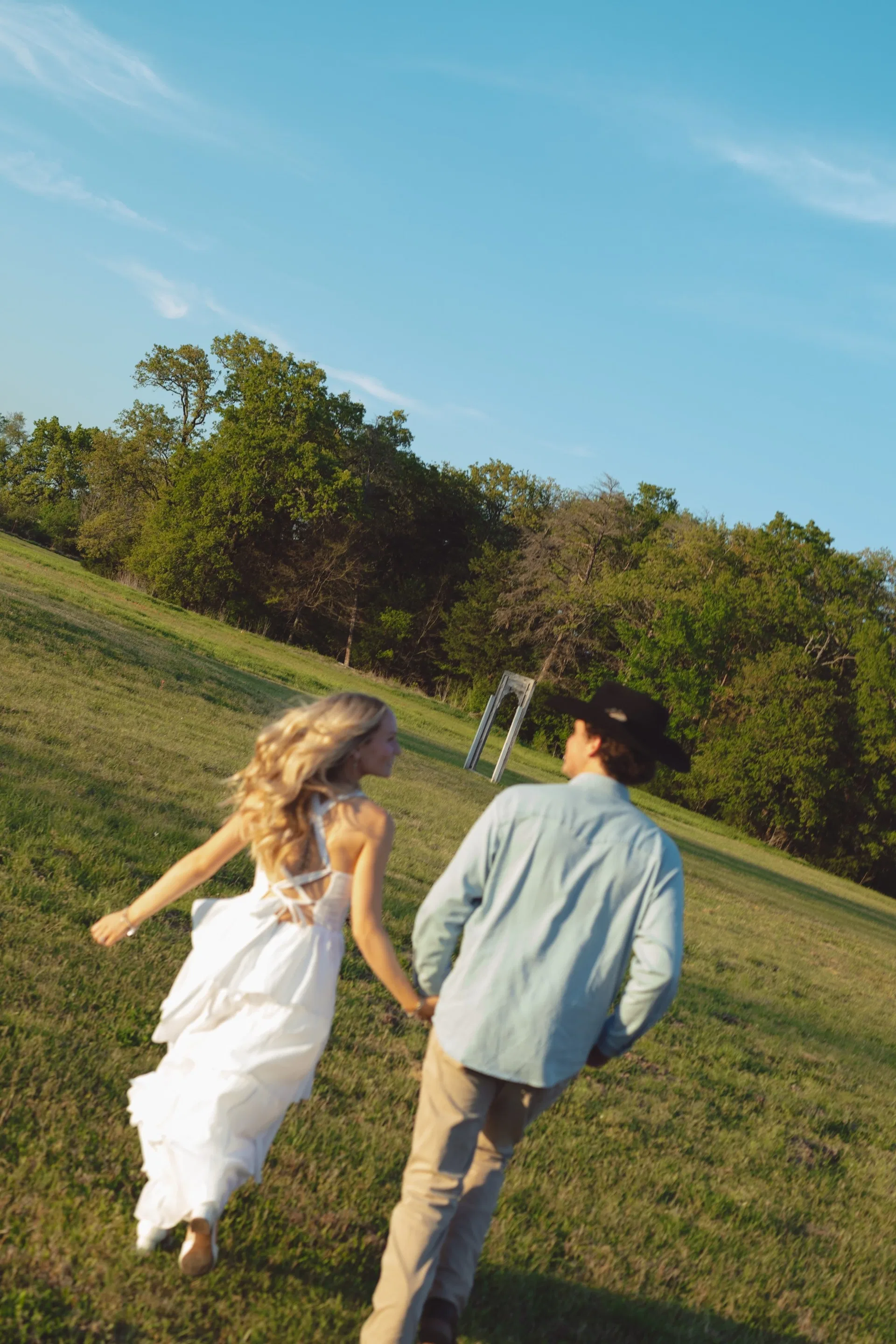 Couple hand in hand crossing a sunlit pasture toward a distant ceremony arch