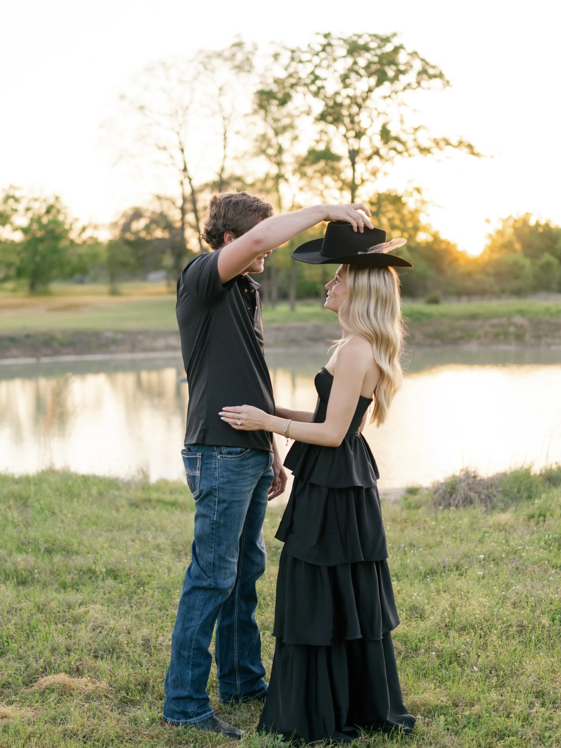 He crowns her with his feathered cowboy hat beside a golden-hour pond