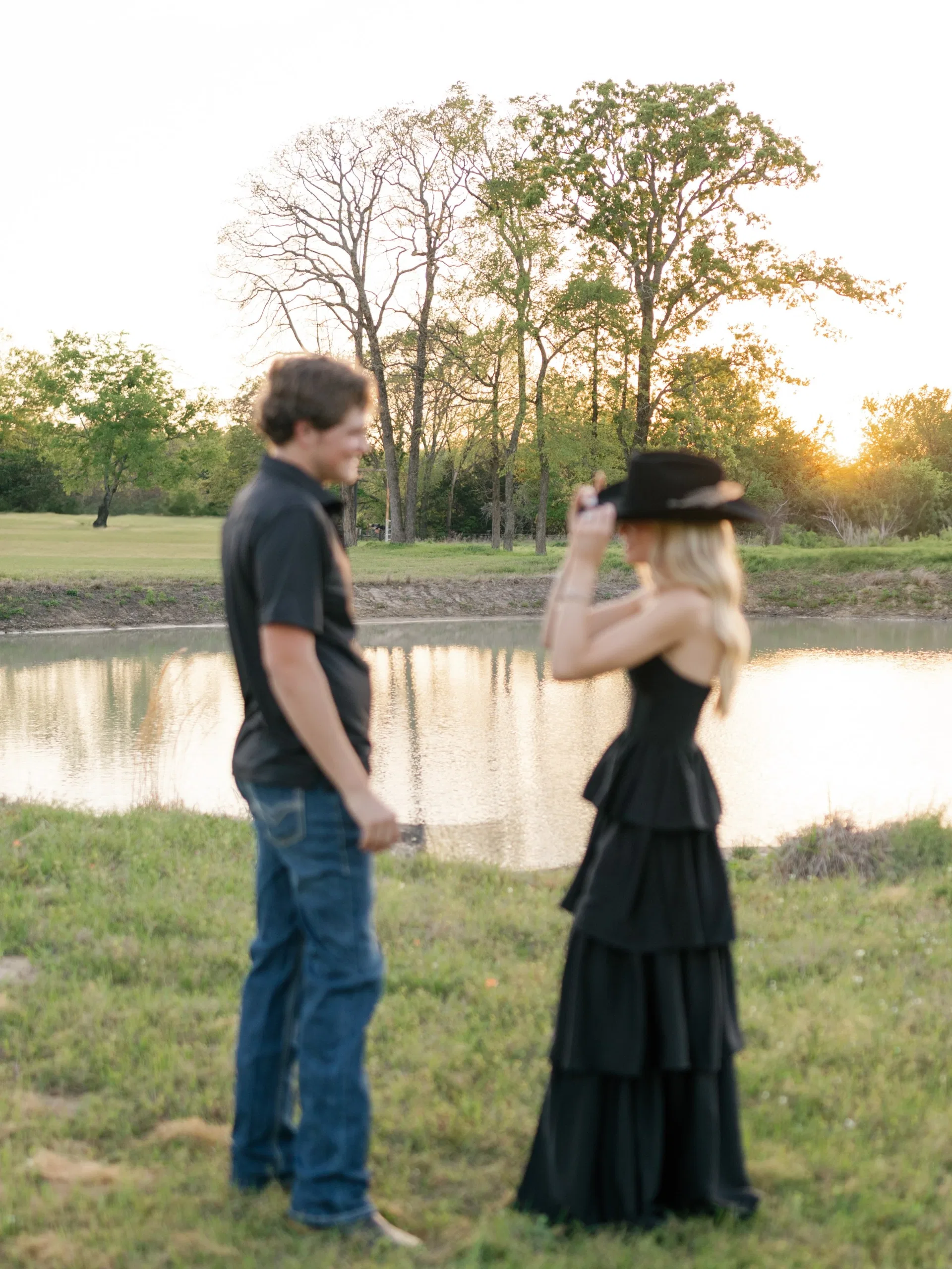 She tips her black cowboy hat toward him beside a sunset pond