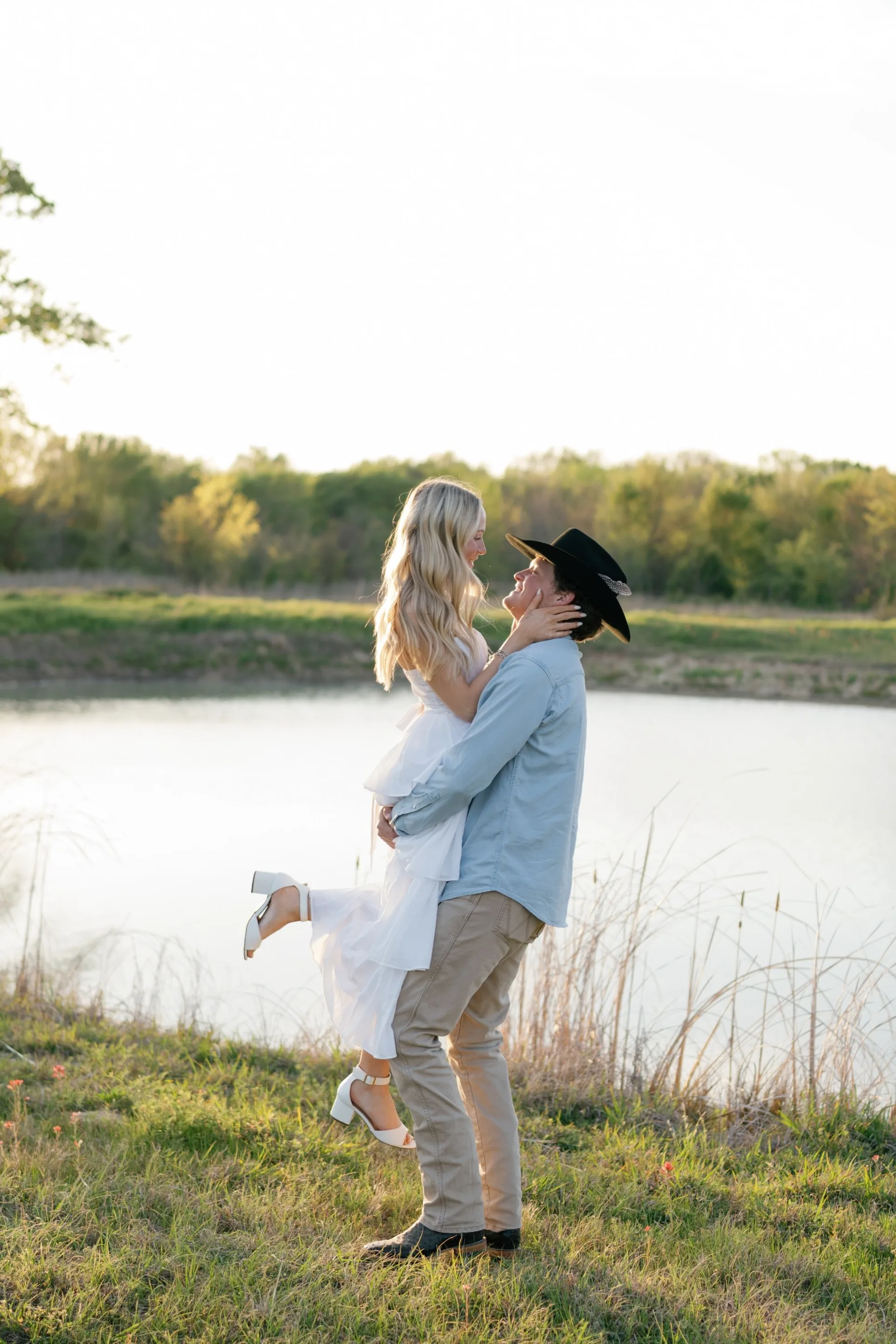 Groom in black cowboy hat lifting his bride beside a Texas pond at golden hour