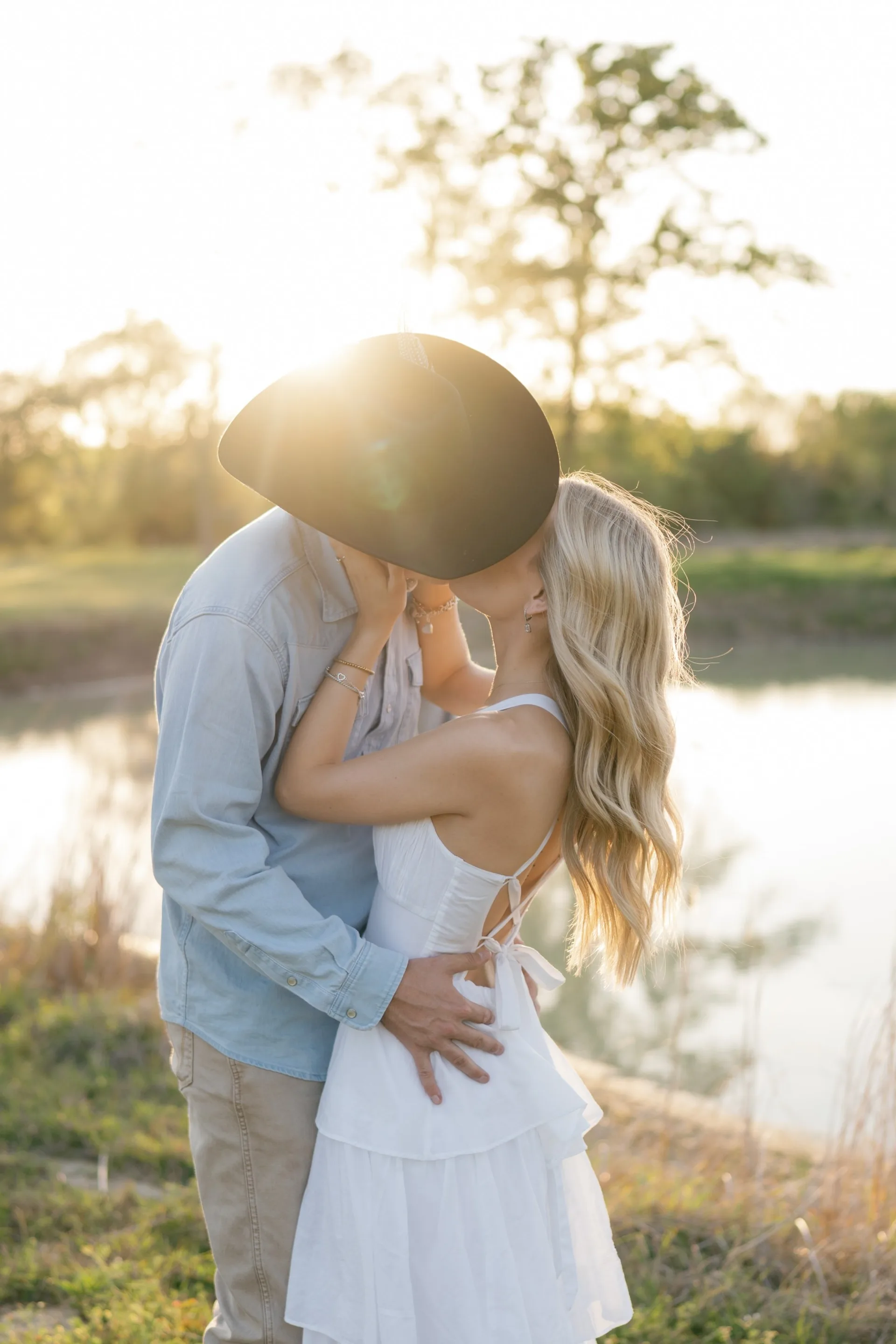 Sunburst flaring behind a cowboy hat as a couple kisses beside a still pond