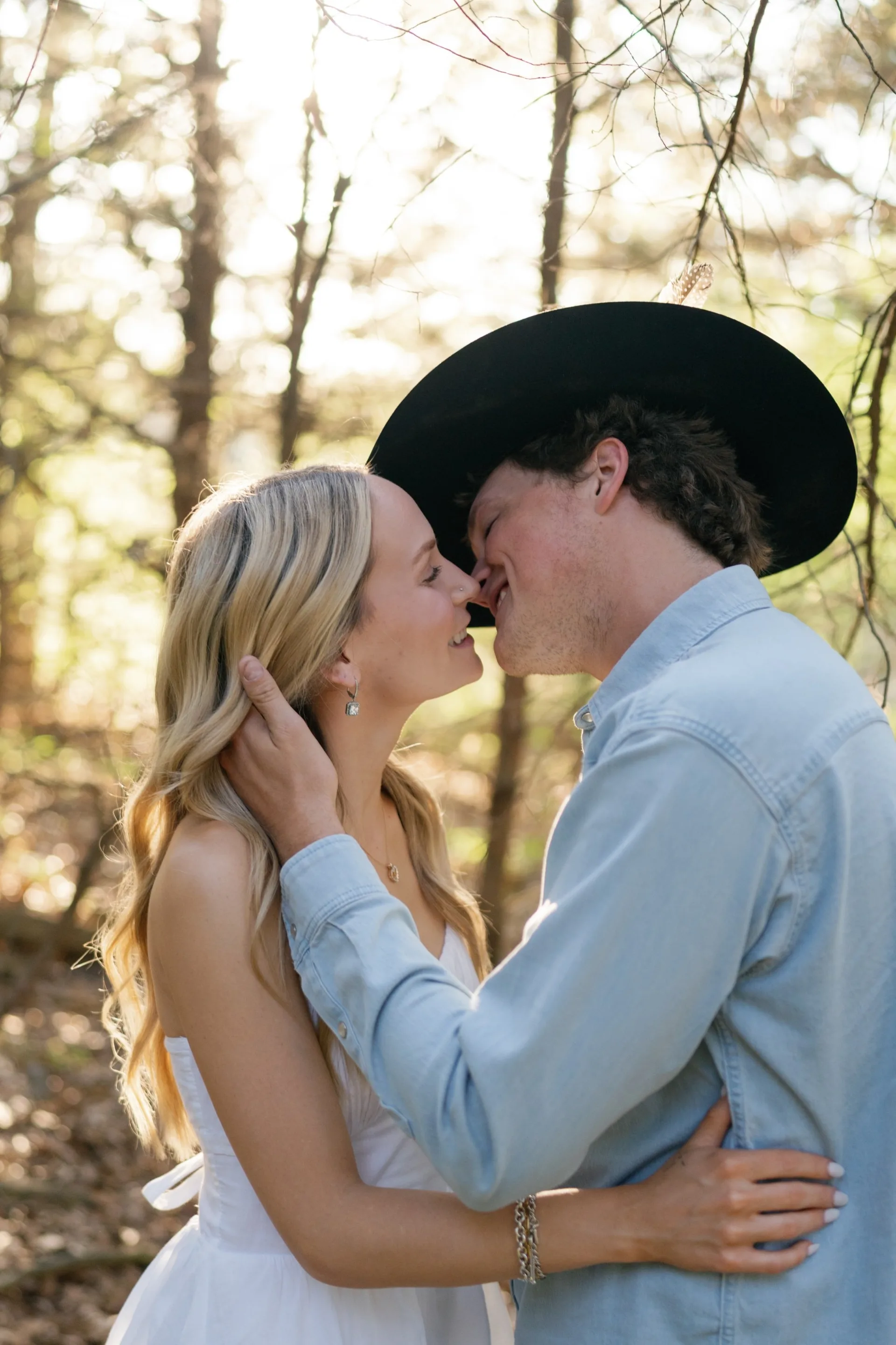 Kiss in a sunlit forest during a Texas engagement session, black hat tipped low
