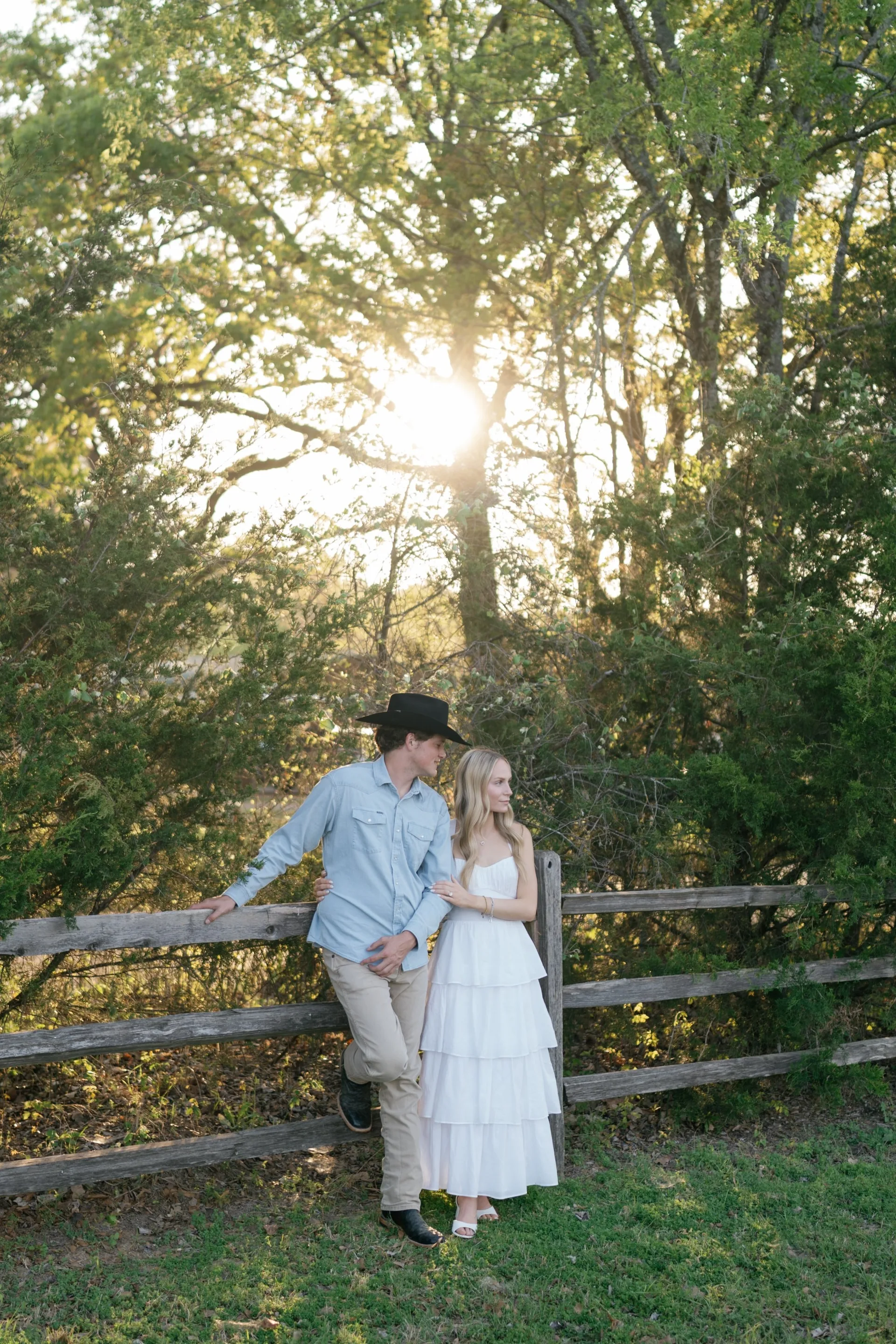 Couple resting against a weathered split-rail fence beneath sunlit country oaks