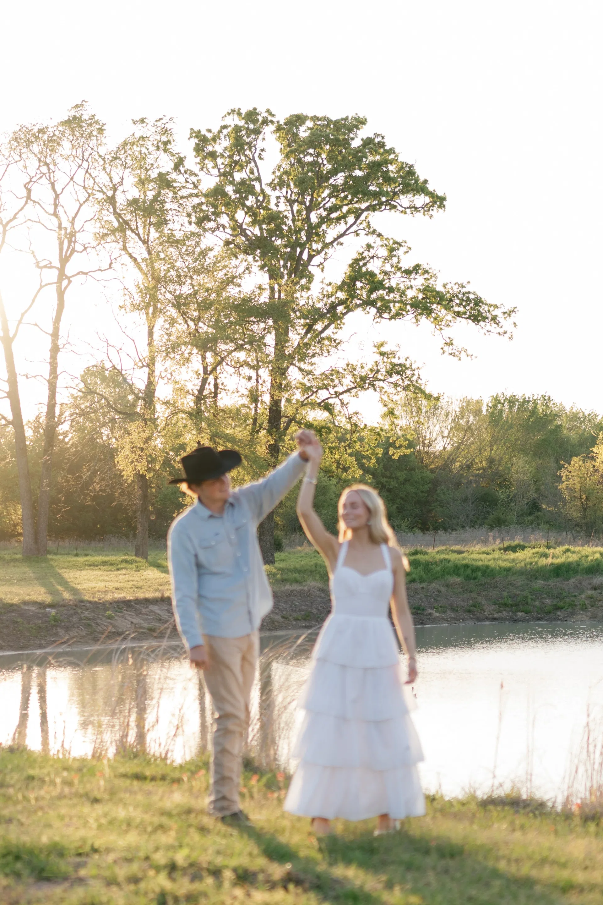 Groom in cowboy hat twirling his bride under raised arms beside a glowing pond