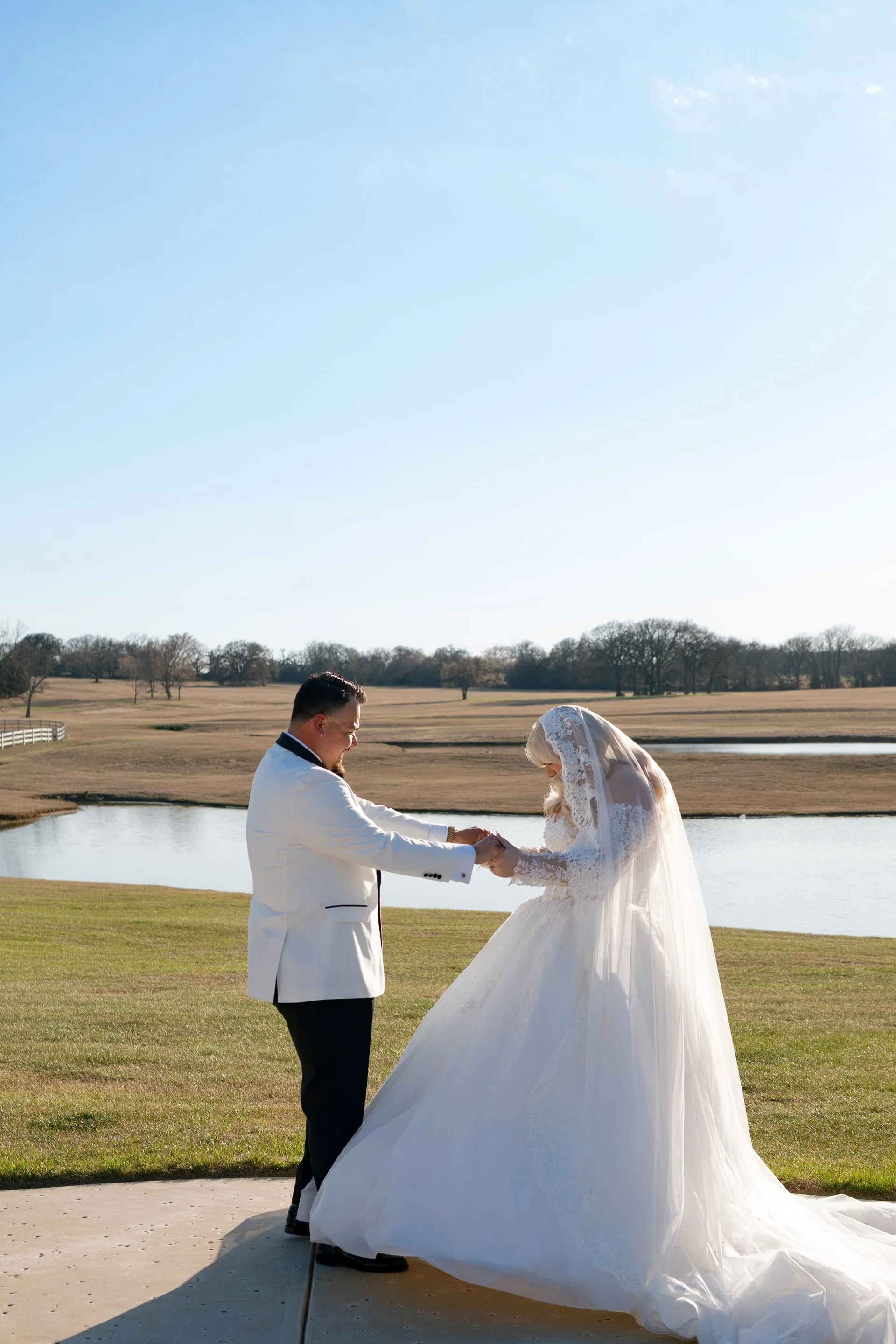 Groom in white tuxedo jacket holding his bride's hands beside a quiet ranch pond