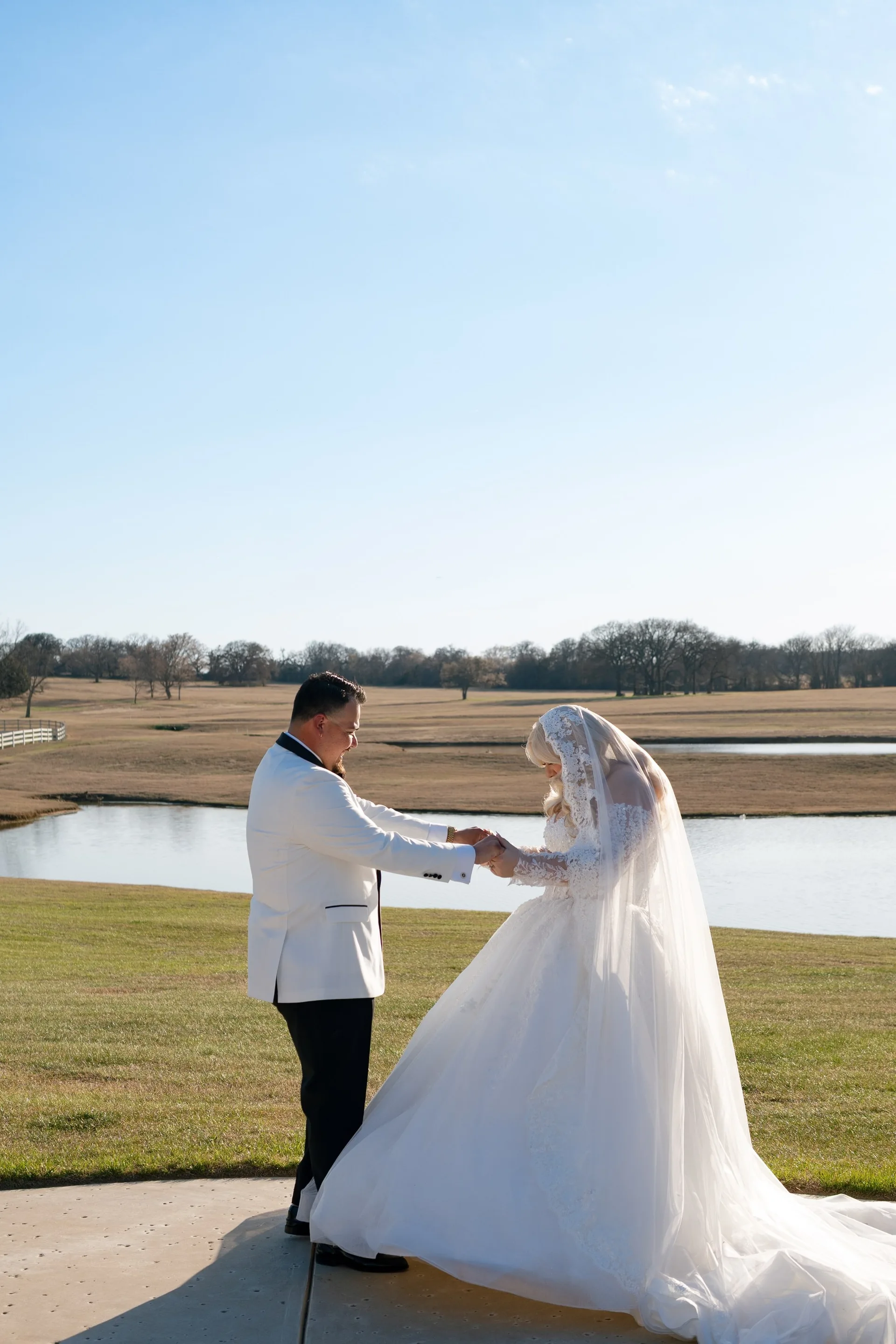 Groom in white dinner jacket holding his bride's hands beside a still ranch pond