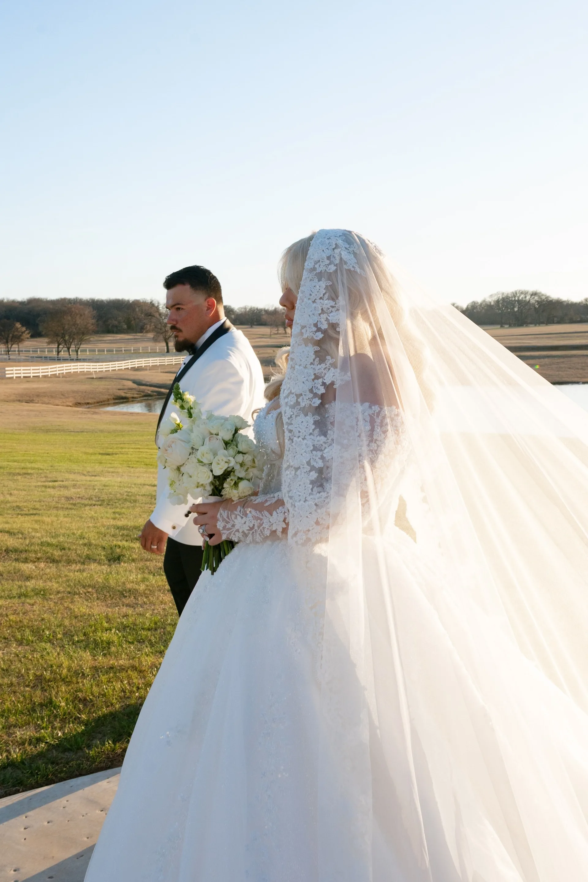 Bride in lace-edged cathedral veil carrying white roses beside her groom on ranch land