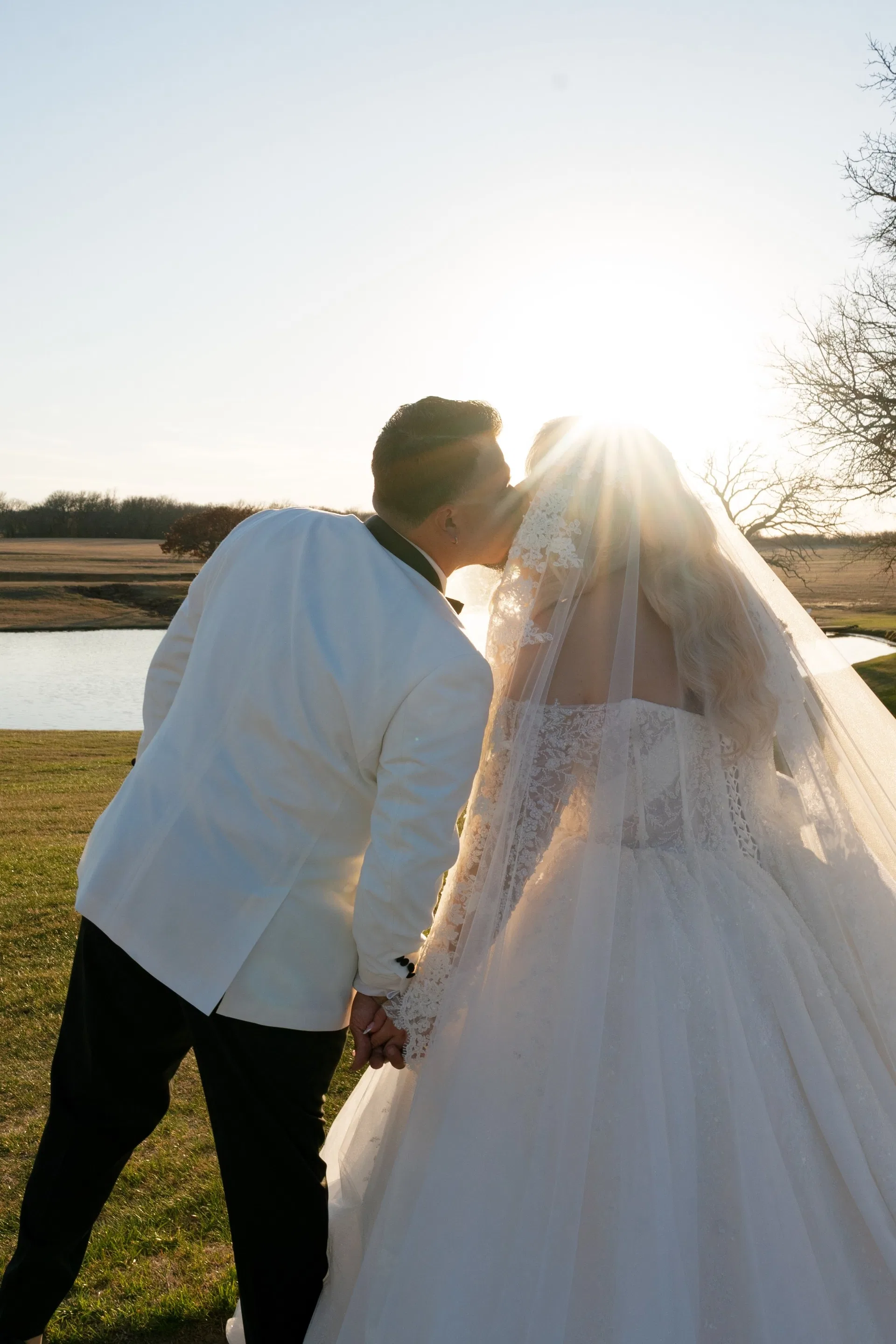 Groom in white dinner jacket kissing his bride beneath a starburst sun by the lake