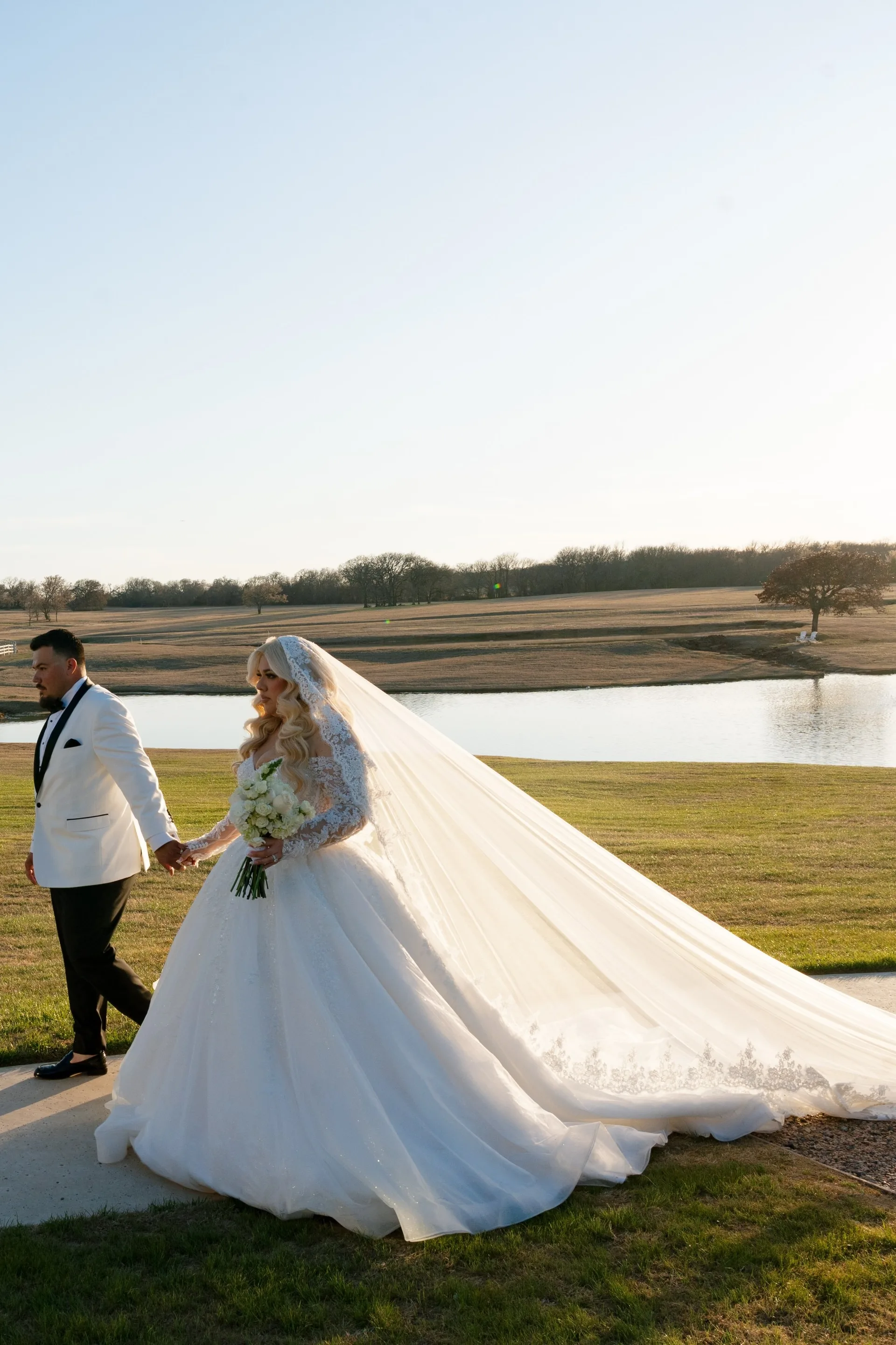 Groom in white dinner jacket leading his bride, cathedral veil trailing across ranch grounds