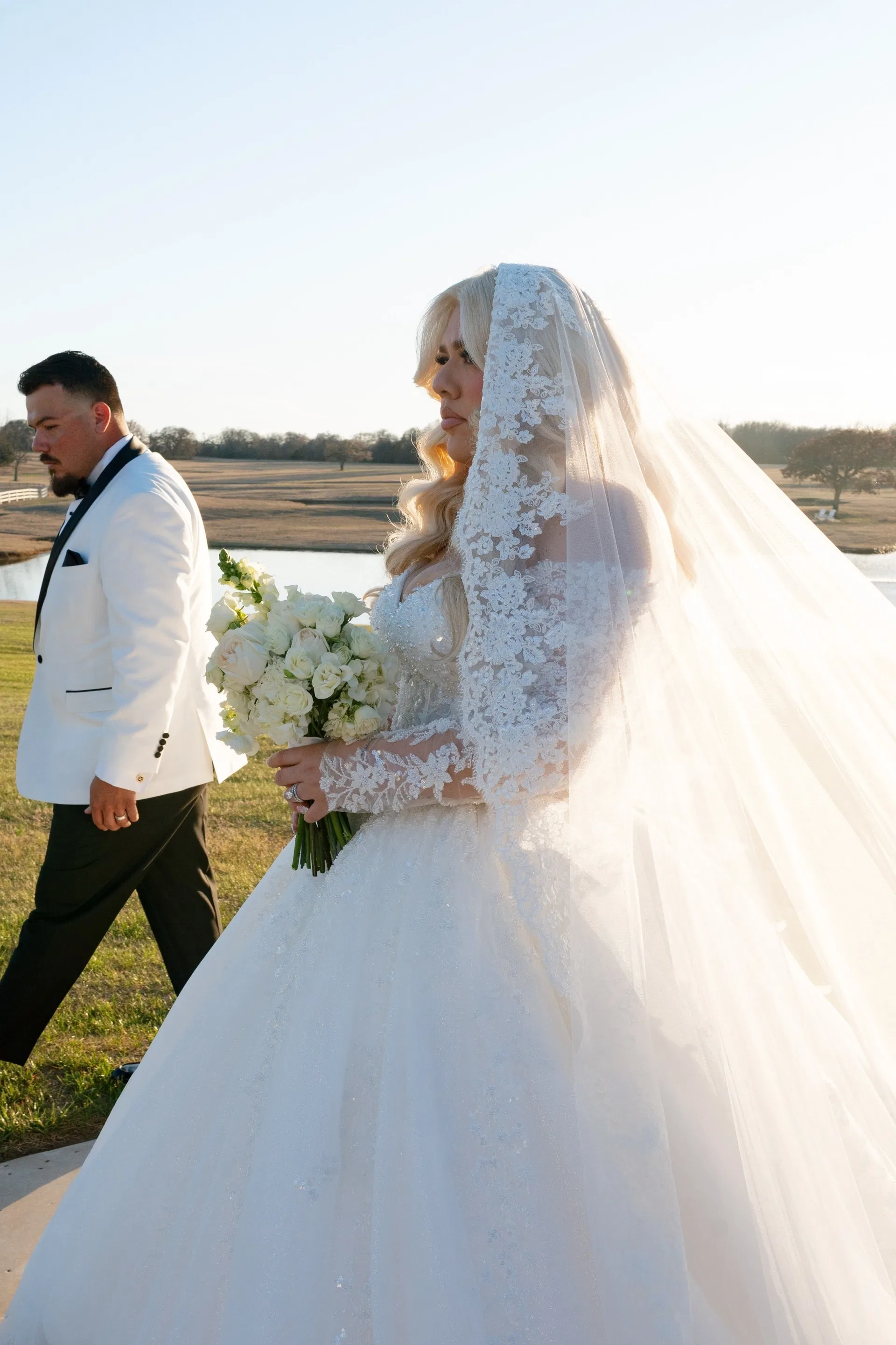 Bride in mantilla veil carrying white roses across a Texas ranch at sunset