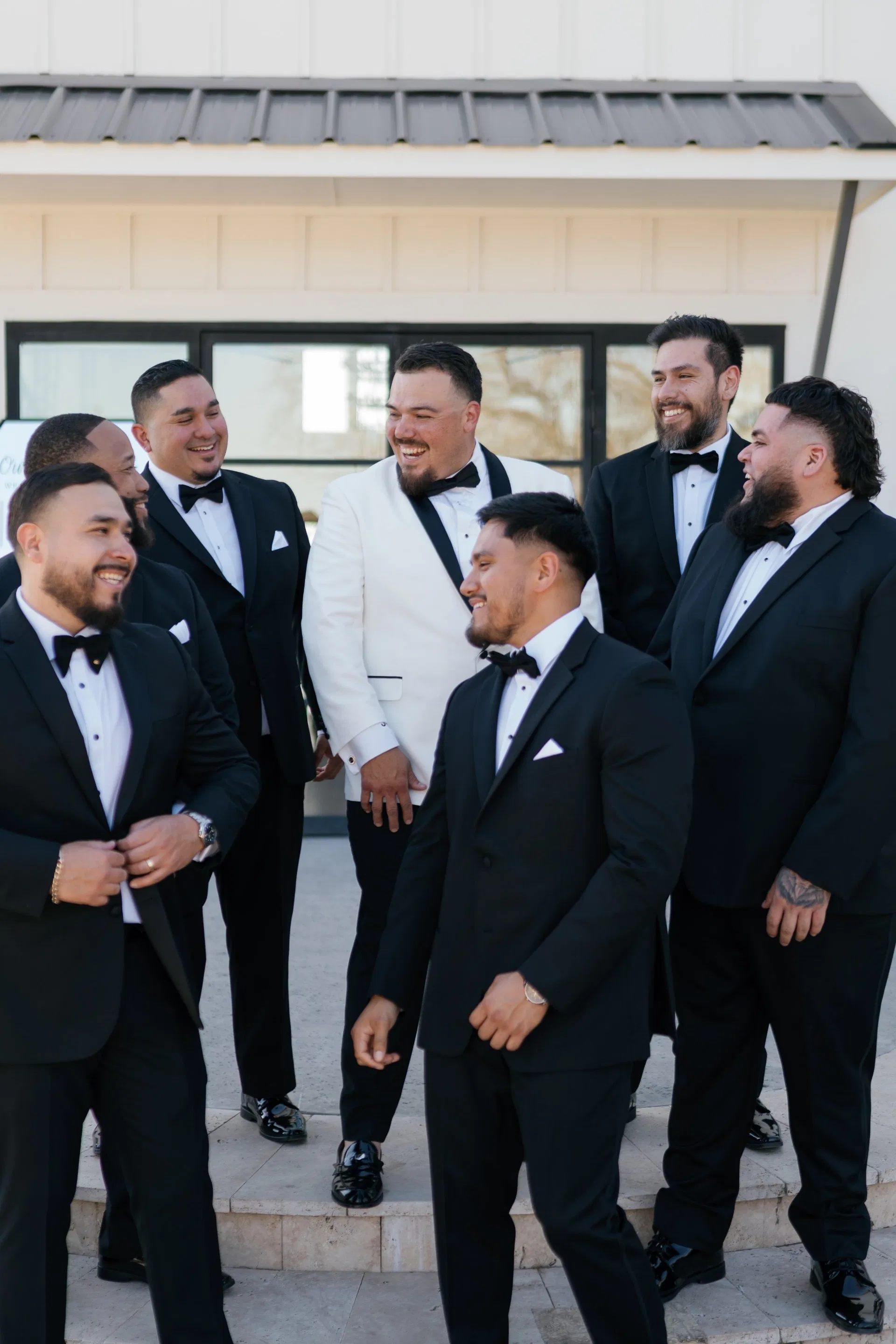 Groom in white jacket laughing with groomsmen outside a modern farmhouse venue