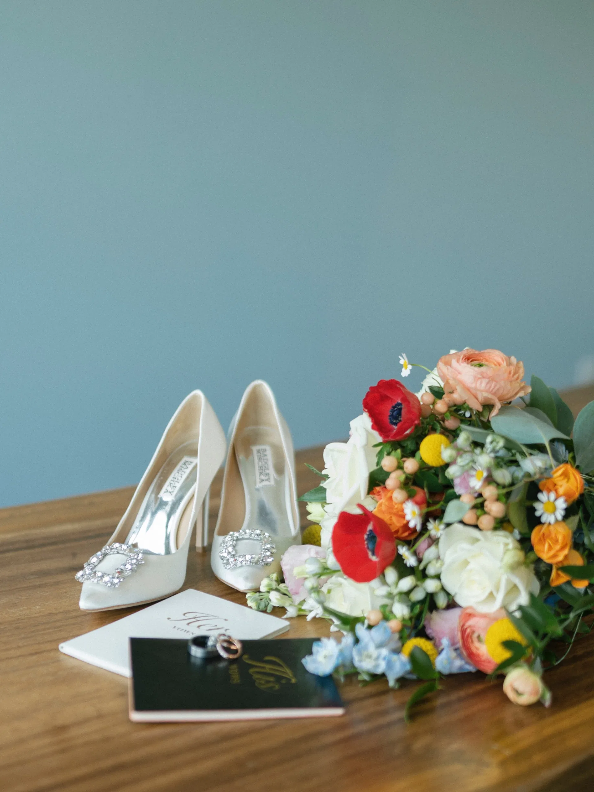 Bridal heels, rings, vow books, and a vivid wildflower bouquet styled on wood