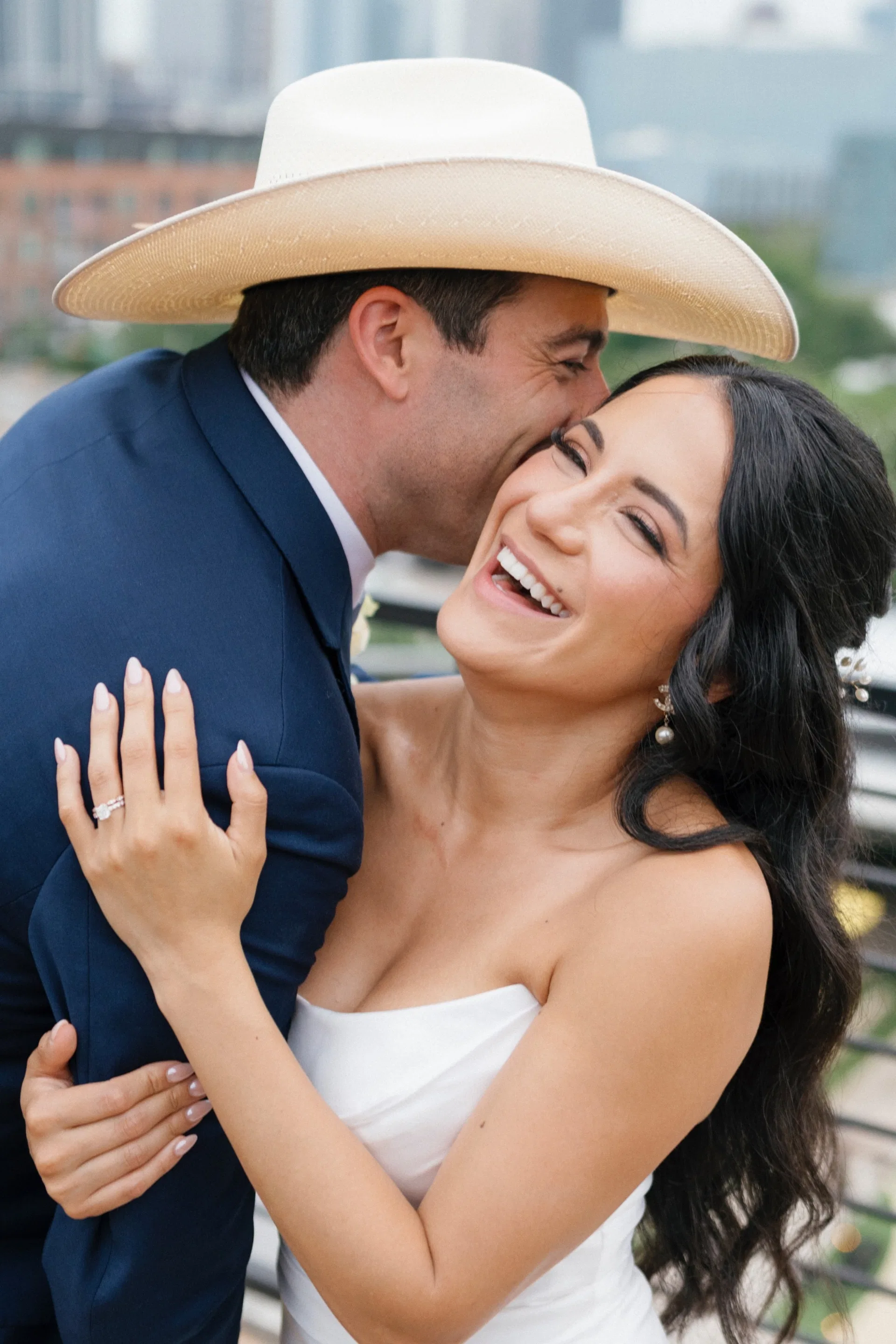 Groom in straw cowboy hat kissing his laughing bride above the Dallas rooftops