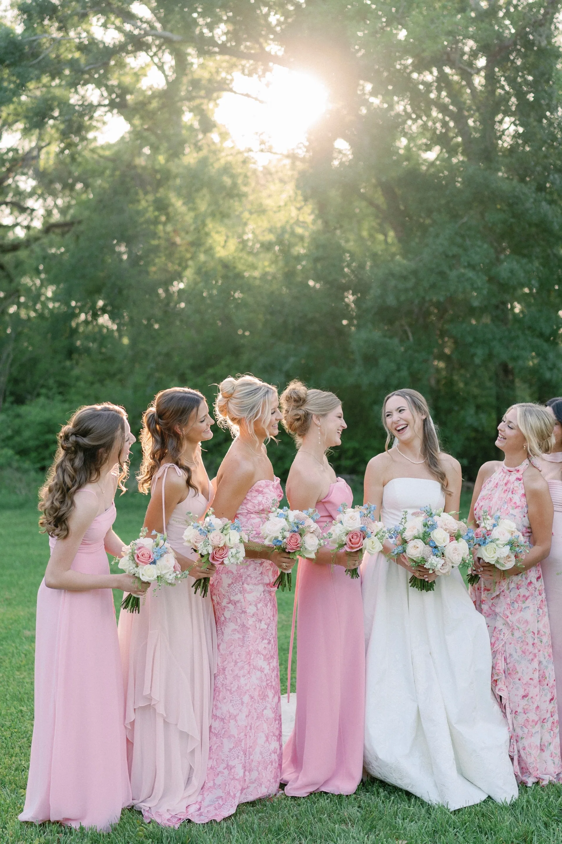 Bride laughing with bridesmaids in blush pink gowns beneath sunlit trees at a garden wedding
