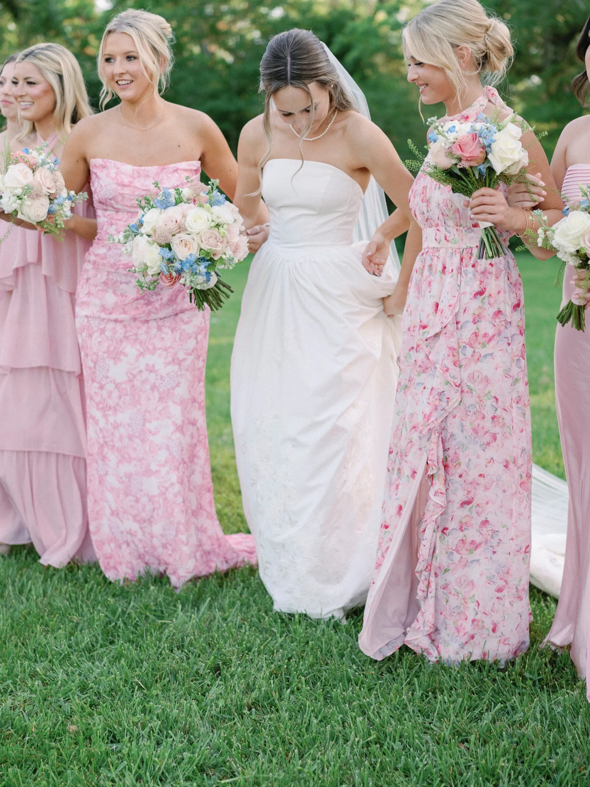 Bride and bridesmaids in shades of pink walking together across a green lawn