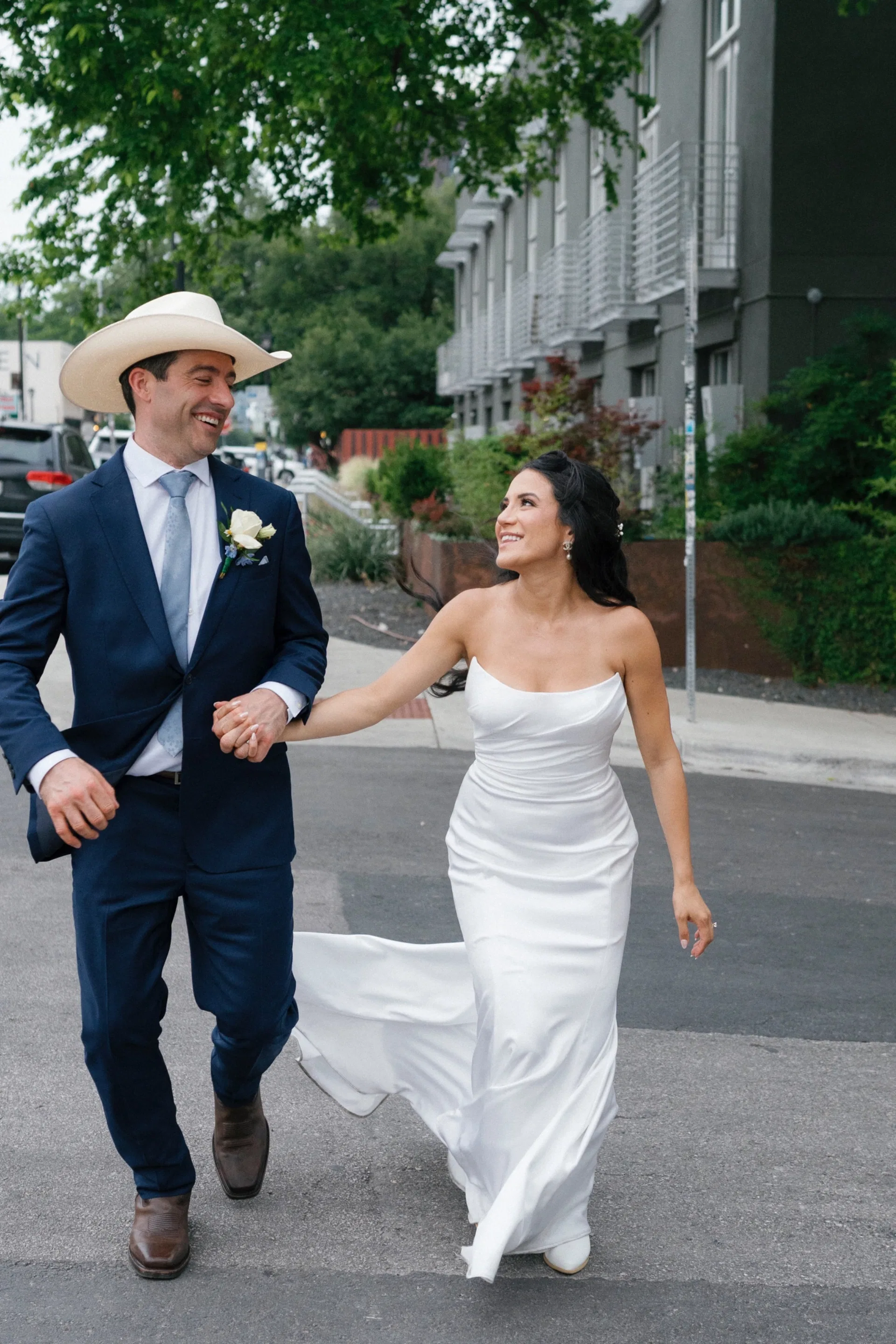 Newlyweds in cowboy hat and strapless gown crossing a Dallas street, laughing