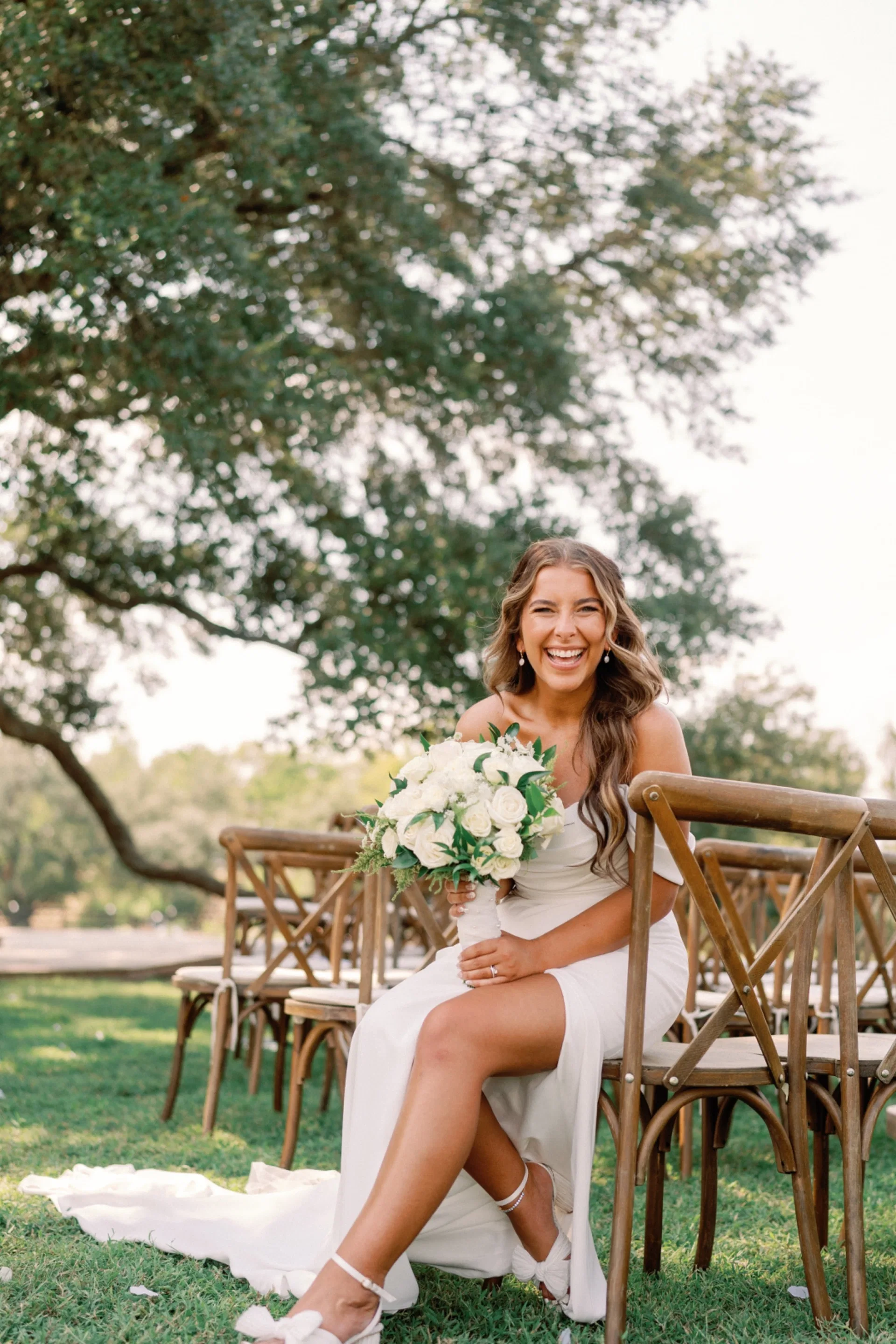 Bride laughing with her white bouquet among crossback chairs beneath a sprawling oak
