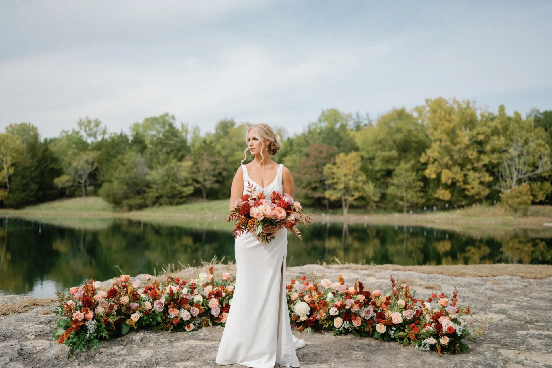 Bride with autumn-toned bouquet standing between ground florals at a lakeside elopement